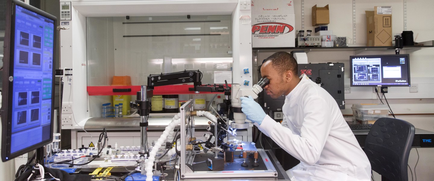 Dark-skinned man in white lab coat looks into microscope in lab setting. 